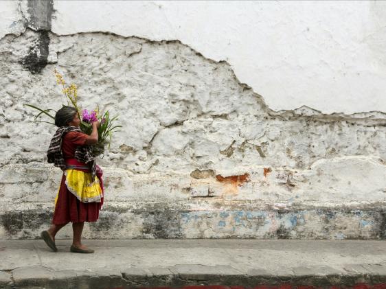 A woman carries a basket of flowers next to the cobblestone streets and crumbling walls of Antigua, Guatemala.